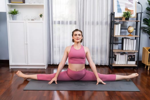 Flexible and dexterity woman in sportswear doing yoga position in meditation posture on exercising mat at home. Healthy gaiety home yoga lifestyle with peaceful mind and serenity.