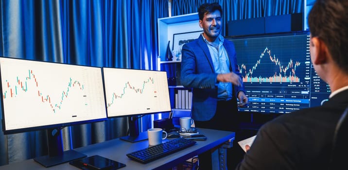 Two stock exchange traders discussing dynamic valued investment rate on monitor at night in panorama photo view. Businessman in stock market presenting economic in neon light at workplace. Sellable.