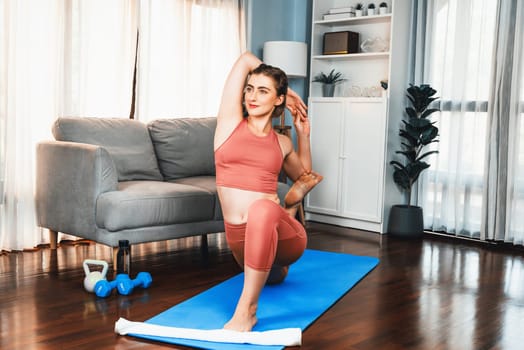 Flexible and dexterity woman in sportswear doing yoga position in meditation posture on exercising mat at home. Healthy gaiety home yoga lifestyle with peaceful mind and serenity.