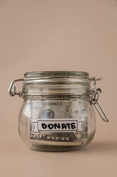 Saving Money In Glass Jar filled with Dollars banknotes. DONATE transcription in front of jar. Managing personal finances extra income for future insecurity. Beige background