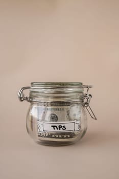Saving Money In Glass Jar filled with Dollars banknotes. TIPS transcription in front of jar. Managing personal finances extra income for future insecurity. Beige background