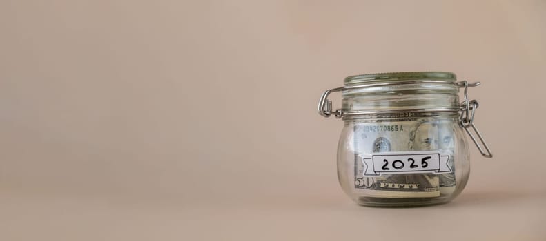 Saving Money In Glass Jar filled with Dollars banknotes. 2025 year transcription in front of jar. Managing personal finances extra income for future insecurity. Beige background