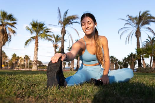 Young woman stretching hamstring sitting on the grass in park during workout. Female stretching after running outdoors. Fitness concept.