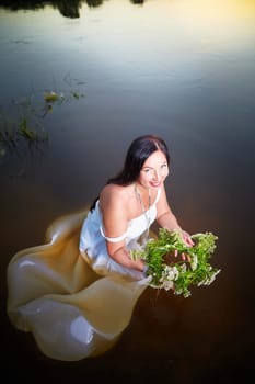 Adult mature brunette woman in a white dress, sundress and a wreath of flowers in summer in water of river or lake in evening at sunset. Celebration of the Slavic pagan holiday of Ivan Kupala