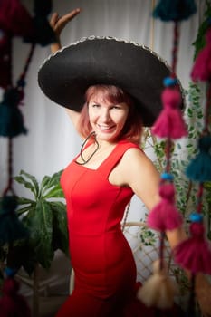 Funny, cute, self-confident adult girl posing in red dress and a huge hat in the room. A middle-aged model at a photo shoot in the studio. Leisure activities