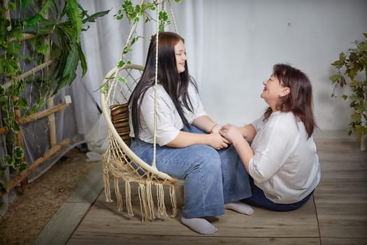 Happy Overweight family with mother and daughter in room on a wicker swing. Middle aged woman and teenager girl having fun, joy and hugging