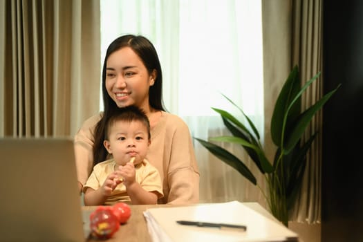 Shot of a young mother having video call with her clients over laptop and carrying her baby on laps.