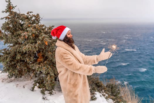 Outdoor winter portrait of happy smiling woman, light faux fur coat holding heart sparkler, posing against sea and snow background.