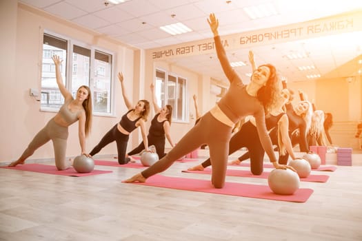 A group of six athletic women doing pilates or yoga on pink mats in front of a window in a beige loft studio interior. Teamwork, good mood and healthy lifestyle concept