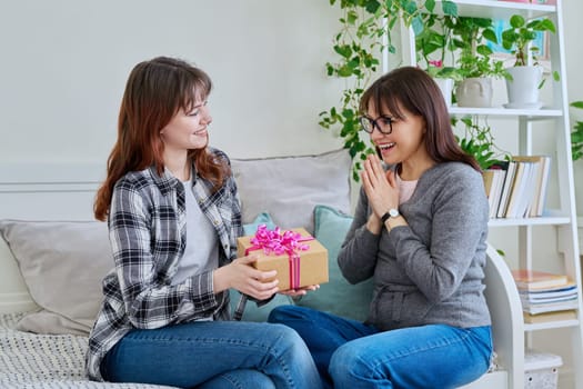 Cheerful teenage daughter congratulating middle aged mother with gift, sitting together on couch at home. Holiday birthday, congratulations, love, mother's day, happiness joy concept