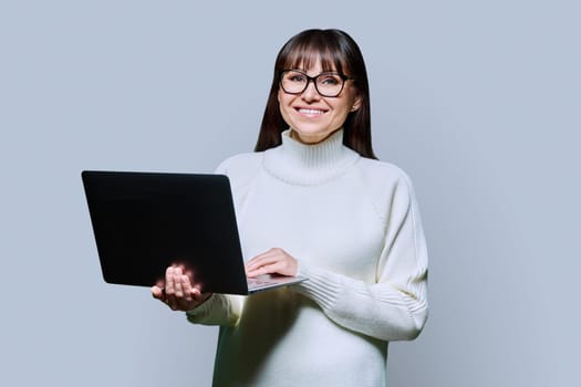Middle-aged beautiful woman holding laptop looking at camera on grey studio background. Smiling happy female in white sweater using computer. Technologies for business work communications leisure