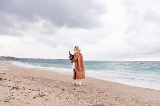 Blond woman Christmas sea. Christmas portrait of a happy woman walking along the beach and holding a Christmas tree in her hands. She is wearing a brown coat and a white suit