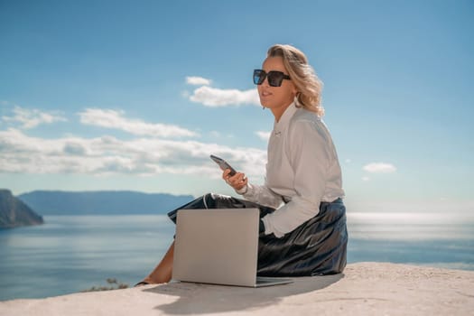 Freelance women sea working on a computer. Pretty middle aged woman with computer and phone outdoors with beautiful sea view. The concept of remote work