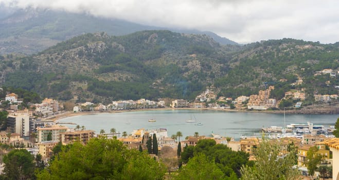 Clouds caress the mountain tops above Port de Soller, a picturesque bay cradling the calm sea and coastal town.