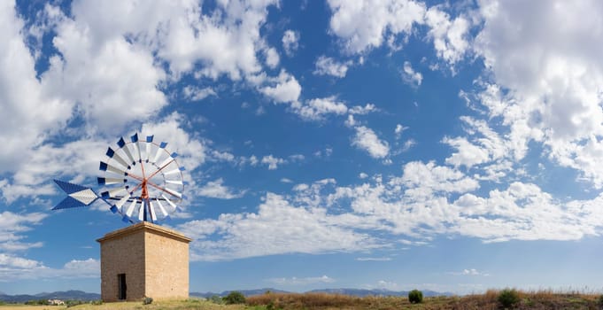 A classic windmill, with striking blue and white blades, graces the rustic landscape under the vast Mediterranean sky.