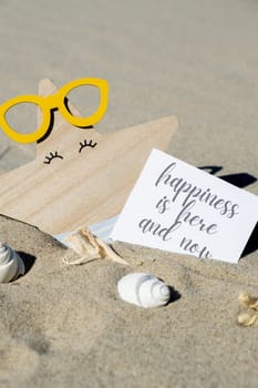 HAPPINESS IS HERE AND NOW text on paper greeting card on background of funny starfish in glasses summer vacation decor. Sandy beach sun coast. Slowing-down, enjoying the moment, good moments, slow life Holiday concept postcard. Getting away Travel Business concept