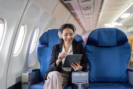 A woman is sitting on a blue airplane seat with a tablet in her hand. She is smiling and she is enjoying her time on the plane