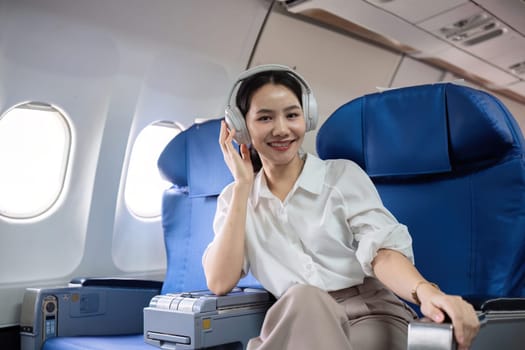 Young woman wearing headphones listening to music during travel, sitting near window in first class on airplane during flight, travel and business concept.