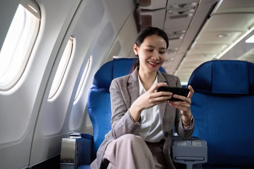 Young Asian woman checks business news on mobile phone, sitting near window in first class on airplane during flight, travel and business concept.