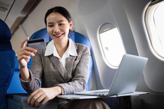 Young Asian woman holding credit card shopping online using website on laptop Sitting near window in first class on airplane during flight, travel and business concept..