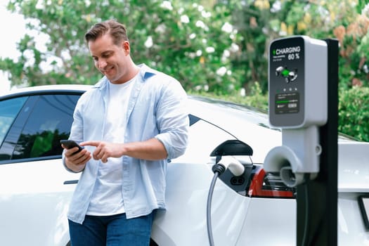 Modern eco-friendly man recharging electric vehicle from EV charging station, using Innovative EV technology utilization for tracking energy usage to optimize battery charging on smartphone.Synchronos