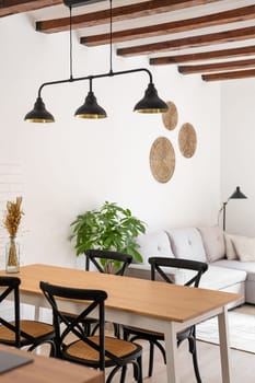 Vertical shot of a kitchen with a wooden table and chairs against the backdrop of a living room with loft lamps and a beamed ceiling. Scandinavian interior concept in a new building.