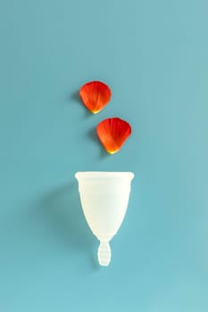 Eco Silicone Reusable Menstrual Cup Isolated on Blue Background with Red Petals as Blood Drops Above. Absorbing Period Flow. Personal Intimate Hygiene. Woman Menstruation Cycle, Gynecology. Vertical.