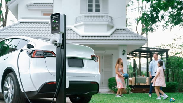 Focus electric vehicle recharge from home charging station on blur background of happy and playful family playing together. EV car using alternative and sustainable energy for better future.Synchronos