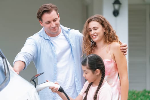 Happy little young girl learn about eco-friendly and energy sustainability as she help her family recharge electric vehicle from home EV charging station. EV car and modern family concept. Synchronos