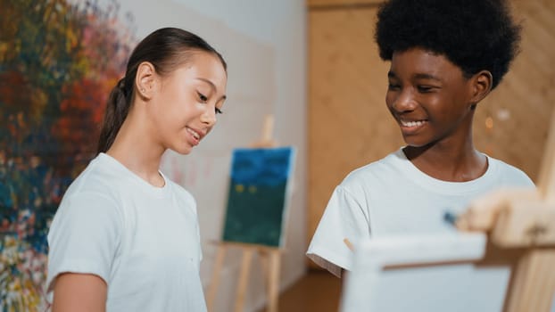 Cute girl looking canvas while boy painting picture in art lesson. Diverse highschool student standing at colorful stained wall while multicultural children attend in creative activity. Edification.