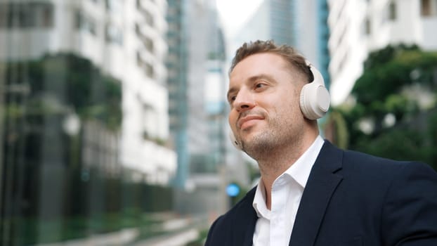 Caucasian businessman listen headphone and dancing to relaxing music with cheerful and motivation. Skilled project manager enjoy to listen relaxed song with blurred background at urban city. Urbane.