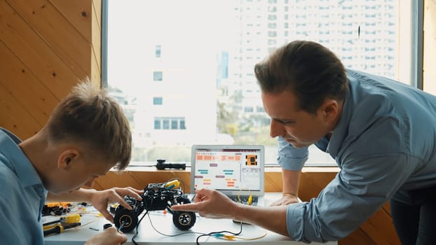 Teacher help student fixing car model and giving advise for programing system or coding prompt while highschool boy listen and use equipment to repair robot at STEM classroom. Back view. Edification.