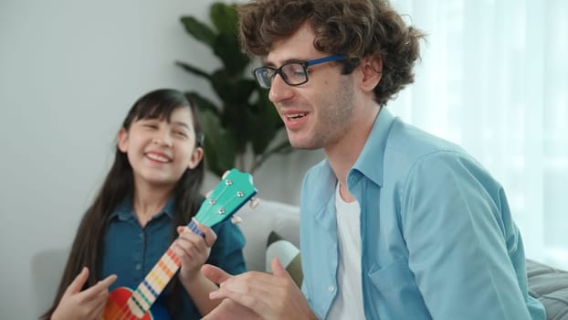 Back view of happy father looking at smart daughter playing ukulele warmly and singing song. Happy dad motivated and encouraged american school girl enjoy playing guitar. Family recreation. Pedagogy.