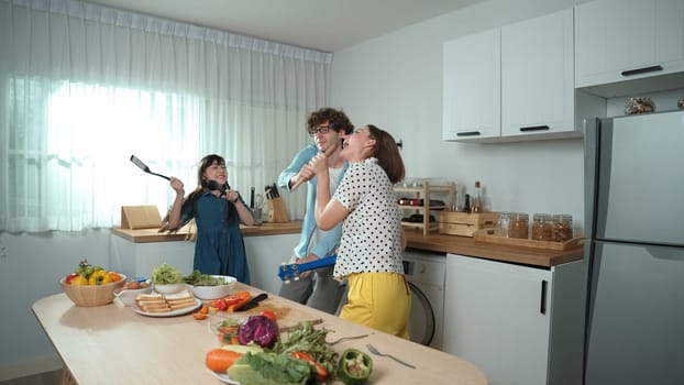 Caucasian skilled father, mother and asian daughter making breakfast while dancing together. Skilled mom playing ukulele while preparing vegetable at modern kitchen. Healthy food concept. Pedagogy.