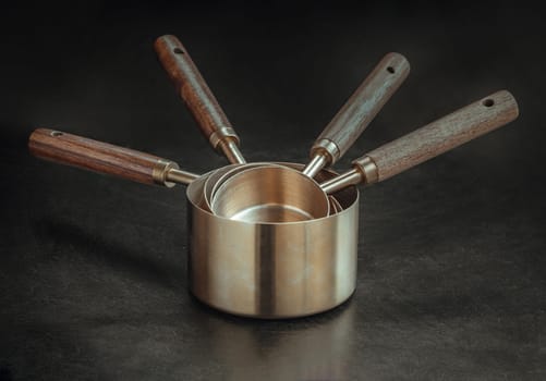 Set of Brass Measuring Cups with Wood Handles with Hanging Hole Design on dark background. Wooden & Brass Measuring Cups & Spoons for Measuring Dry and Liquid Ingredients, Kitchen tool, Copy space, Selective Focus.