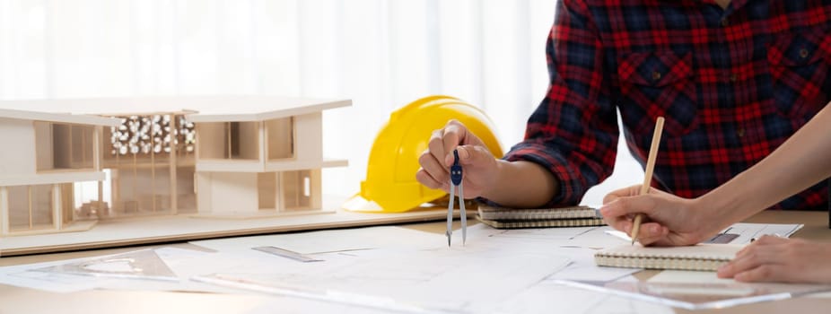 Cropped image of cooperative architect team decide and work together on meeting table with house model, safety helmet and architectural plan scatter around. Closeup. Focus on hand. Burgeoning.