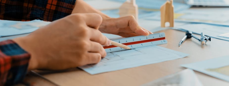 Closeup of architect engineer hand using ruler to mature and draw a blueprint on meeting table with wooden block, pencil and blueprint scatter around at architectural modern office. Delineation.