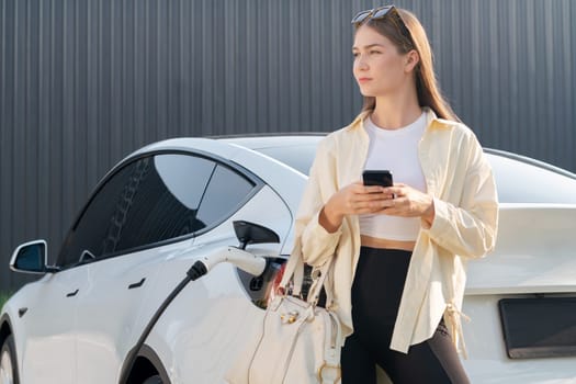 Young woman holding shopping bag and use smartphone to pay for electricity for recharging EV car battery from charging station at city mall parking lot. Modern woman go shopping by eco car. Expedient