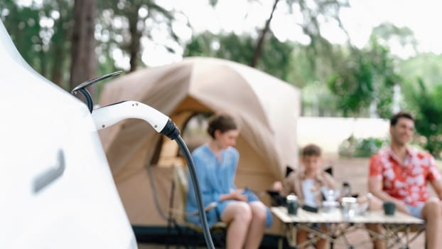 Outdoor adventure and family vacation camping in nature travel by eco friendly car for sustainable future. Lovely sit on trunk, charging EV car with EV charging station in campsite. Panorama Perpetual