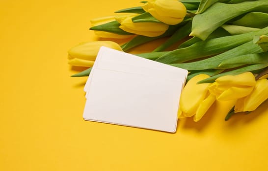 Blank paper white business card and a bouquet of yellow tulips on a yellow background, close up