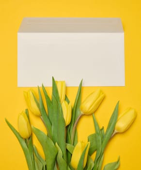 Envelope and bouquet of blooming yellow tulips with green leaves on a yellow background, top view