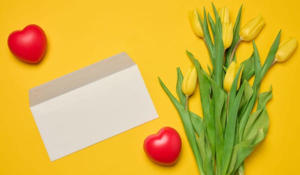 Envelope and red heart and bouquet of blooming tulips with green leaves on a yellow background, top view