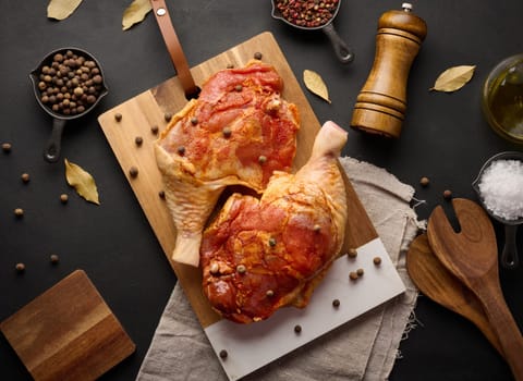 Raw chicken drumsticks seasoned on a wooden board, accompanied by salt and peppercorns, viewed from the top. Black table