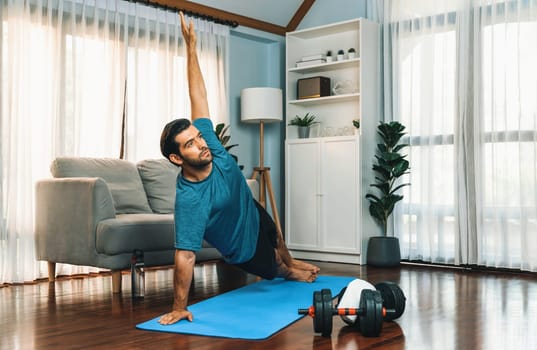 Flexible and dexterity man in sportswear doing yoga position in meditation posture on exercising mat at home. Healthy gaiety home yoga lifestyle with peaceful mind and serenity.