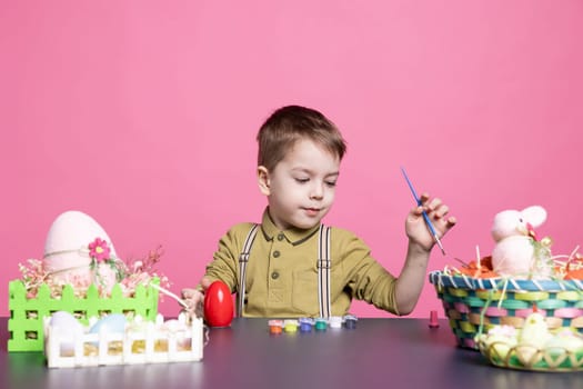 Lovely youngster with a smile crafting eggs and decorations for the Easter festivities, using art supplies and watercolor paint. Little pleased boy feeling excited about painting activity.