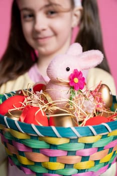 Young cheerful kid holding a colorful decorations basket on camera, showing painted eggs and a stuffed pink rabbit as festive arrangement. Cute girl smiling in studio with adorable decor. Close up.