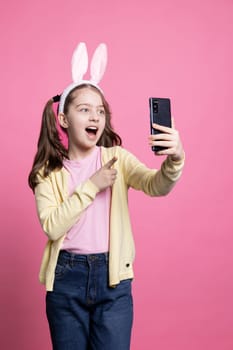 Adorable confident girl calling her friends on videocall online, chatting about easter celebration holiday while she wears bunny ears. Young joyful child holding smartphone and waving at webcam.