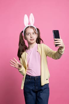 Confident young kid uses phone network to meet on videocall with her school friends against pink background, joyful toddler kid on internet conference. Cheerful small girl with bunny ears.