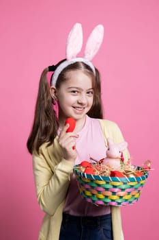 Smiling young girl with bunny ears showing red eggs and a basket filled with colorful items and a rabbit in studio. Enthusiastic little kid feeling happy with easter decorations for festive event.