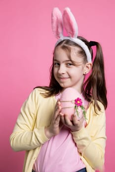 Playful schoolgirl with bunny ears holding a pink egg on camera, presenting her handmade decorations for easter celebration. Young adorable child with pigtails feeling pleased with ornament.